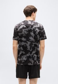 Μαύρο κοντομάνικο πουκάμισο με καμουφλάζ σχέδιο, στρογγυλή λαιμόκοψη και μαλακό ύφασμα, συνδυασμένο με μαύρα σορτς, σε θέα από πίσω.