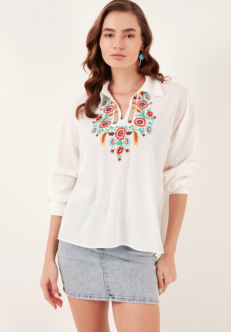Camicia bianca con ricami floreali colorati sul colletto e sul petto, chiusura a bottoni, vestibilità rilassata, abbinata a una minigonna di jeans.