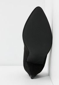 Chaussure à talon haut en daim noir avec un bout pointu et une semelle noire texturée, présentant un design élégant et un matériau supérieur lisse.