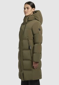 Giacca imbottita verde oliva con colletto alto, cappuccio e tasche con zip. Presenta un design trapuntato e tessuto resistente.