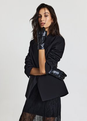 Gants en cuir noir portés avec un blazer noir sur mesure et une robe en dentelle, présentant un design élégant, une texture mate et une coupe ajustée.