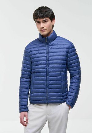 Mladi moški, oblečen v modro prešito jakno z visokim ovratnikom in bele hlače, stoji pred enobarvnim svetlim ozadjem.