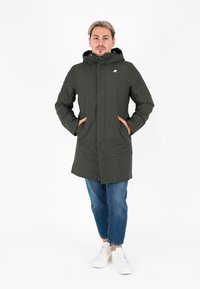 Giacca verde con cappuccio, apertura con zip, due tasche laterali e logo bianco. Indossata sopra jeans blu e sneakers bianche su uno sfondo bianco.