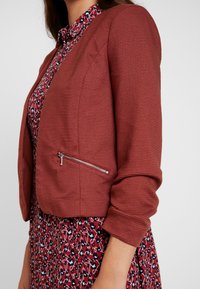 Rust-colored, ribbed jacket with a fitted design and silver zipper pocket. Worn over a floral patterned dress in red, white, and navy.