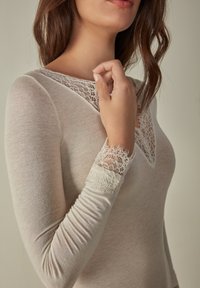 Top de manga larga beige con un escote en V de encaje y acentos de encaje en los puños. Textura suave, diseño ajustado y superposiciones de encaje detalladas.