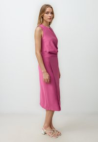 Tailliertes ärmelloses Kleid in leuchtendem Pink, mit drapiertem Oberteil und asymmetrischem Saum. Kombiniert mit hellen, schlichten Absatzsandalen.