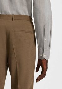 Pantaloni marroni con una superficie liscia, dotati di una sola tasca posteriore. Camicia button-up grigia chiara con un motivo sottile e bottoni ai polsini.