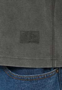 Γκρι ύφασμα με υφή βάφλας και ετικέτα λογότυπου Vans. Εκτεθειμένες ραφές και ίσιο τελείωμα, σε συνδυασμό με μπλε τζιν παντελόνια που εμφανίζουν πορτοκαλί ραφές.