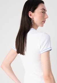 Witte geribde t-shirt met lichtblauwe gestreepte mouwen. Model heeft lang haar en draagt zilveren hangoorbellen. Schoon, minimalistisch ontwerp.