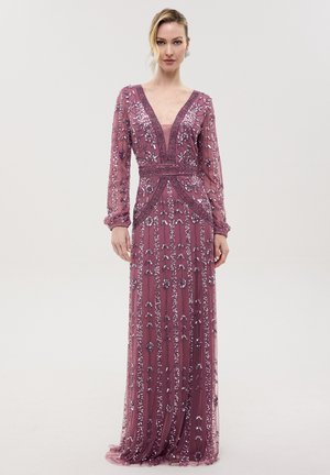 Femme portant une robe longue mauve à manches longues avec des motifs floraux et géométriques en sequins, se tenant devant un fond clair uni.