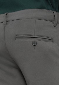 Šedé kalhoty s hladkým, elastickým materiálem. Zadní kapsa s horizontálním otvorem a jedním černým knoflíkem.