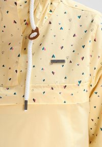 Gul hoodie med små flerfärgade hjärtan, vit dragkedja med brun stopp, och en metallmärke på bröstet.