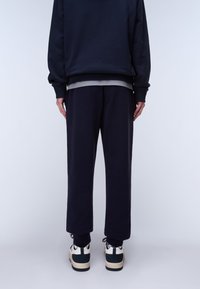 Person steht mit dem Rücken zur Kamera und trägt schwarze Jogginghosen, einen schwarzen Pullover, ein graues Unterhemd und weiße Turnschuhe mit schwarzen Akzenten vor einem schlichten Hintergrund.