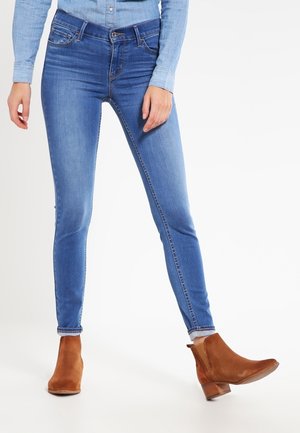 Femme portant un jean skinny bleu, une chemise bleu clair boutonnée et des bottines marron, debout sur un fond blanc.