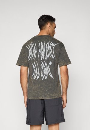 T-shirt à manches courtes, vert délavé avec une texture usée et un texte graphique blanc dans le dos. Associé à un short noir.