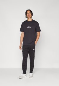 Juodas medvilnės marškinėliai su baltu "adidas" logotipu, derinami su juodomis kargo kelnėmis, turinčiomis kišenes ir šoninį logotipą. Pilkos sportinės bateliai.