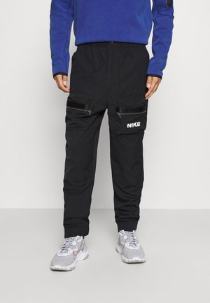 Mężczyzna w czarnych spodniach cargo Nike z kieszeniami na zamek i szarych butach sportowych Nike, stojący na jednolitym jasnym tle.