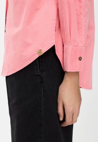 Rosa Cordhemd mit langen Ärmeln, versehen mit geknöpften Manschetten und einem Seitenschlitz. Getragen über schwarzen Jeanshosen, mit sichtbarem Etikett am Saum.