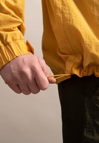 Geel lichtgewicht jack met elastische manchetten, geplooide onderrand en verstelbare koord in de hand. Subtiele textuur zichtbaar op het oppervlak van de stof.