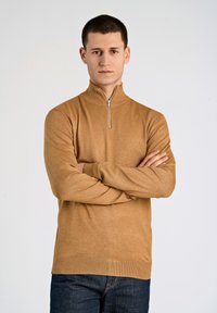 Maglione zip-up beige realizzato in morbido tessuto, con colletto alto e polsini a costine, abbinato a jeans scuri. Design semplice e classico.