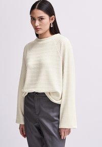 Cremefarbener, strukturiertes Strickpullover mit Rundhalsausschnitt und langen, weiten Ärmeln. Kombiniert mit dunkelgrauen Hosen, die einen geraden Schnitt und eine Knopfverschluss haben.