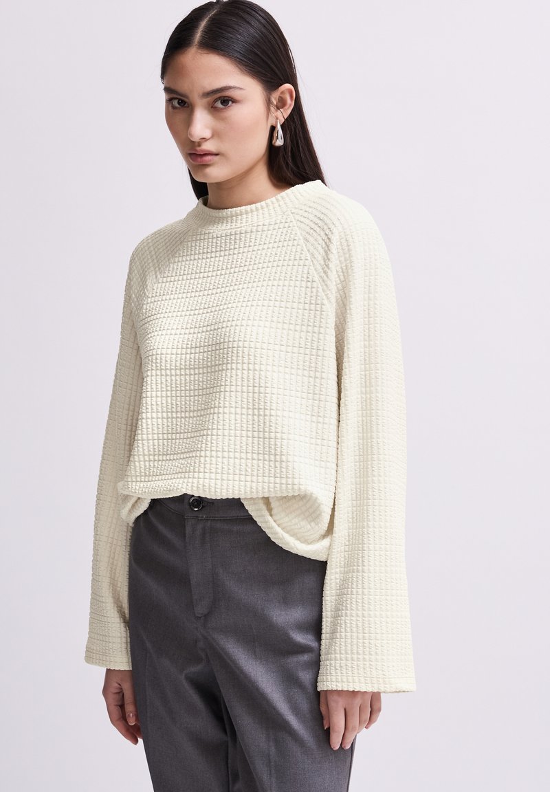 Cremefarbener, strukturiertes Strickpullover mit Rundhalsausschnitt und langen, weiten Ärmeln. Kombiniert mit dunkelgrauen Hosen, die einen geraden Schnitt und eine Knopfverschluss haben.