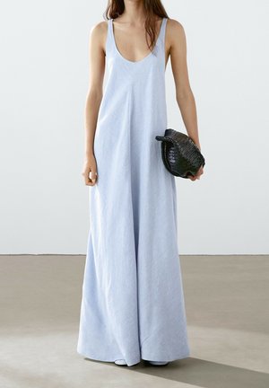 Femme portant une robe longue sans manches bleu clair tenant une pochette noire tissée devant un fond uni.