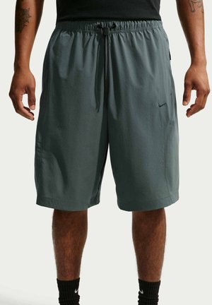 Mand iført mørkegrå sweatshorts med snøre og sort Nike-logo, sorte sokker og sort skjorte, står med armene afslappet ned langs siden.