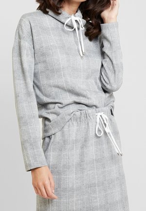 Kobieta ubrana w jasnoszary, w kratę, długi rękaw i dopasowaną spódnicę z białymi troczkami na szyi i w talii.