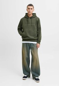 Olivgrüner Kapuzenpullover mit Fronttasche und Kordeln, mit weißem Markentext. Tragen über lässigen, hellblauen Jeans mit weitem Bein und marineblauen Sneakers.