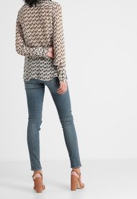 Blusa trasparente e stampata con maniche lunghe nei toni beige e marroni, abbinata a jeans aderenti blu e sandali con tacco marroni.