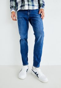 Jeans de mezclilla azul con un corte recto, combinados con zapatillas blancas con detalles en azul marino. El modelo lleva una camisa de cuadros de manga larga.