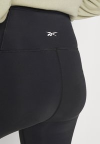 Leggings pretos de cintura alta feitos de um material liso e elástico. Apresentam costuras flatlock e um pequeno logótipo branco perto da cintura.
