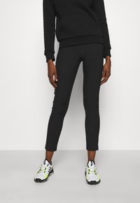 Schwarze, körperbetonte Leggings mit glatter Textur, kombiniert mit weißen und neonakzentuierten Sneakers, präsentiert vor einem schlichten weißen Hintergrund.