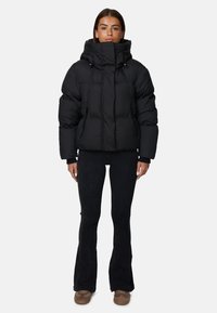 Giacca a vento nera con colletto alto e cappuccio, caratterizzata da una silhouette ampia, grandi camere d'aria e due tasche laterali. Indossata con pantaloni svasati scuri.