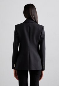 Blazer nero su misura con spalle pronunciate, schiena strutturata e tre accenti di bottoni metallici su ciascun polsino. Tessuto liscio con un design elegante.