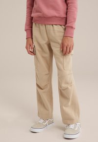 Beige kravas bikses ar taisnu kāju jaukumu, ar sānu kabatām, izgatavotas no izturīgas kokvilnas auduma, kombinētas ar baltām sporta kurpēm.