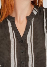 Ciemnobrązowa bluzka z pionowymi białymi paskami, teksturowanym materiałem, kołnierzem mandaryńskim oraz czarnymi guzikami z przodu.