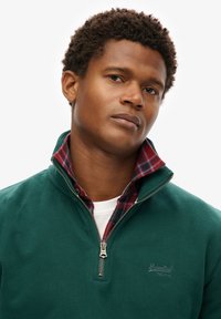 Sudadera verde con cremallera y cuello alto, superpuesta a una camisa de cuadros en rojo y azul. Presenta un logo en el pecho y detalles en la cremallera.