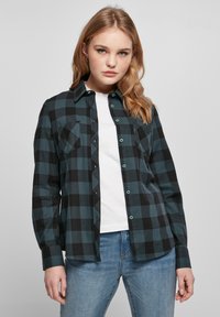 Schwarzes und türkis kariertes Flanellhemd mit Knopfleiste, Brusttaschen und langen Ärmeln, getragen über einem weißen T-Shirt, kombiniert mit hellen Jeans.