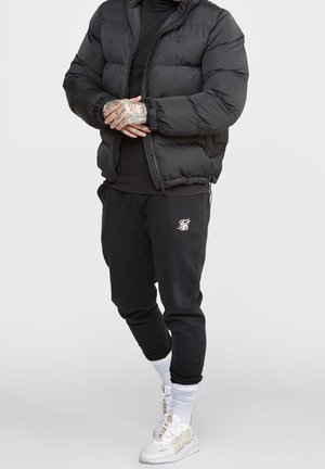 Mand iført sort dunjakke, sorte joggingbukser med hvidt logo, hvide sokker og hvide høje sneakers med beige bogstaver.