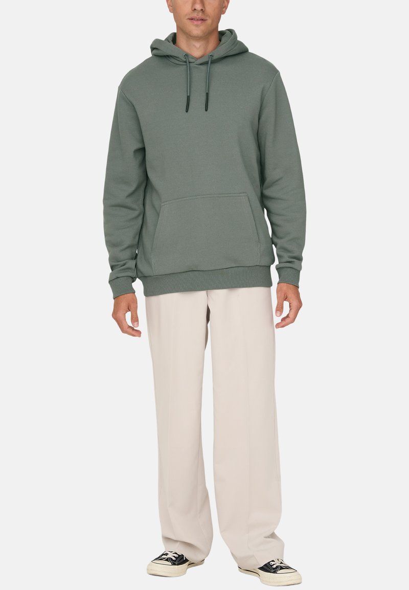 Grøn hoodie med en kengurulomme, ribbede ærmer og kant; parret med beige vidde bukser og sorte sneakers.