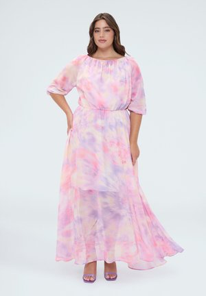 Frau in einem fließenden, pastellrosa und lila Batik-Maxikleid mit Dreiviertelärmeln und violetten offenen High Heels, steht mit einer Hand in der Hüfte.