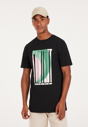 Sort t-shirt med hvid tekst "PROTEST" og farverig grafik af cirkler i pink og grøn. Model bærer en lys beige kasket.