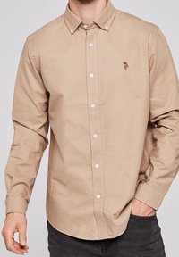 Beige katoenen overhemd met lange mouwen, voorzien van een klein geborduurd detail op de borst en witte knopen. Soepele textuur.