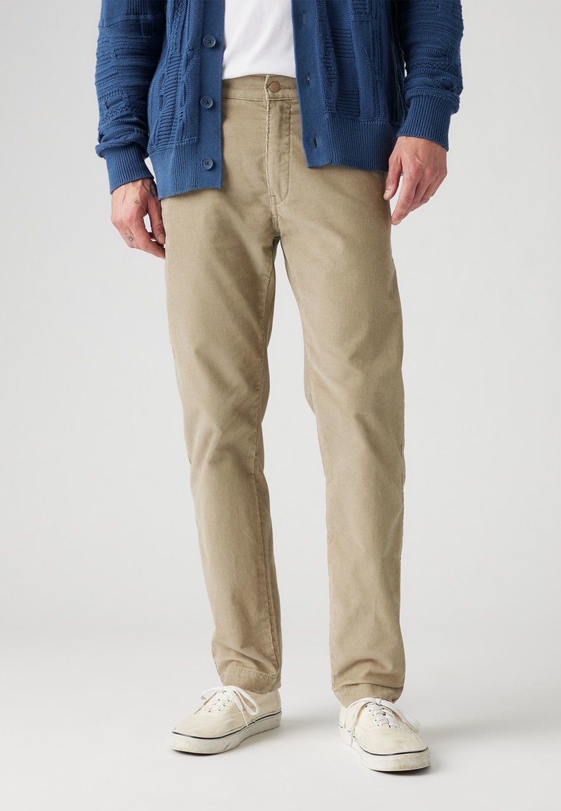 Beige bomullbyxor med rak passform, tillsammans med en strukturerad blå cardigan och vita sneakers, framför en neutral bakgrund.