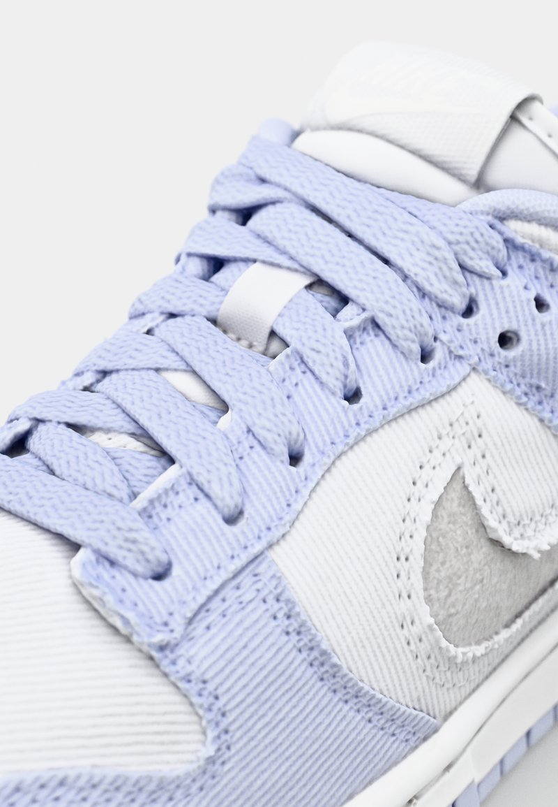Zapatilla blanca con acentos en azul claro, tejido texturizado, costuras detalladas y logo prominente en la lengüeta y el lateral. Cintas planas sobre un cuello acolchado.