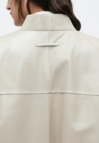 Žena v béžovej koženej bunde s rebrovaným golierom, zobrazená zozadu a z boku, zdôrazňujúca švy a textúru bundy.