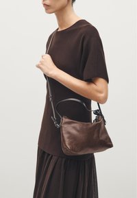 Brown leather handbag with a rounded shape and adjustable strap, held by a person wearing a dark brown top and skirt. Smooth texture, minimalist design.