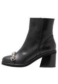 Bota de tobillo de cuero negro con tacón grueso y detalle de cadena plateada en la parte delantera. Textura suave y diseño minimalista y elegante.